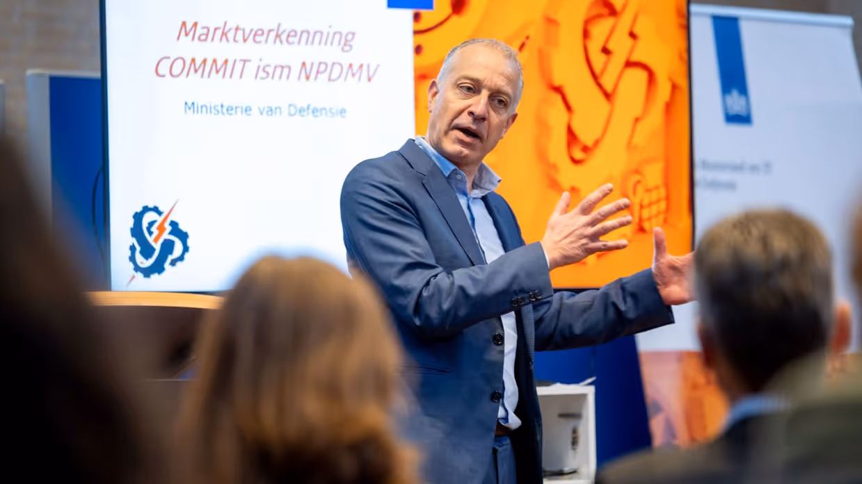 Een man in pak spreekt voor een groep mensen en maakt daarbij gebruik van zijn handen. 