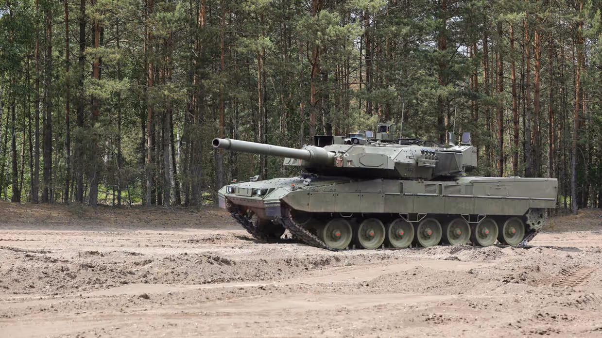 Een rijdende Leopard 2A8-tank rijdt door een oefenterrein met een zanderige ondergrond. Achter het voertuig staan bomen.&nbsp;