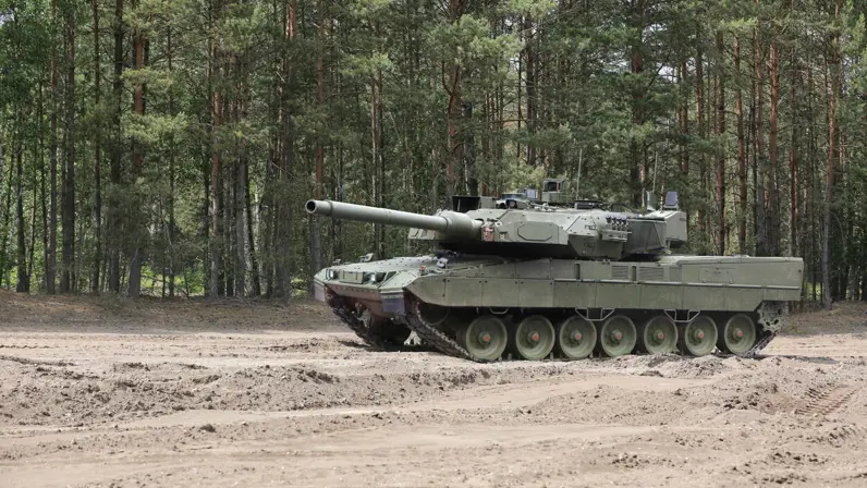 Een rijdende Leopard 2A8-tank rijdt door een oefenterrein met een zanderige ondergrond. Achter het voertuig staan bomen.&nbsp;