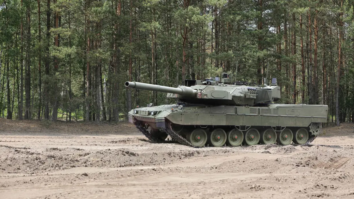 Een rijdende Leopard 2A8-tank rijdt door een oefenterrein met een zanderige ondergrond. Achter het voertuig staan bomen.&nbsp;