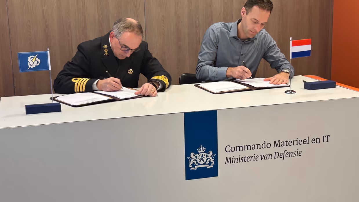 2 heren, van wie 1 in militair DT, achter een desk met pen in de hand voor een ondertekening.
