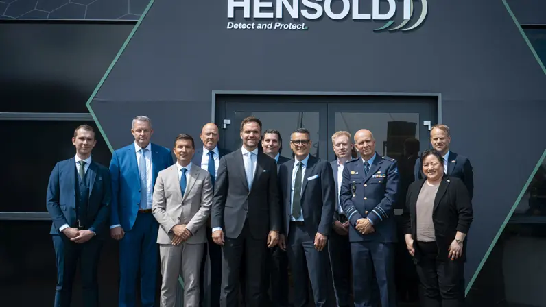 Een groep militairen en burgers poseert voor een donkere muur. Bovenaan de tekst ‘Hensoldt’.
