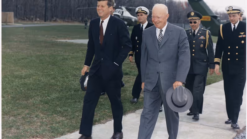 Twee mannen in pak lopen glimlachend over een pad: de jongere draagt donker pak, de oudere grijze jas en hoed, met militairen en helikopter op de achtergrond. 