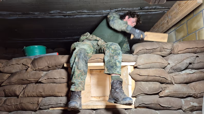 Een persoon in camouflagekledij leunt tegen zandzakken, met zijn voeten op een houten kist, in een schuilplaats.
