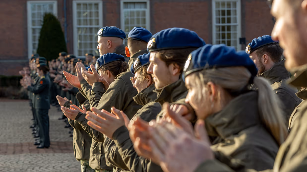  Links: Een groep mensen in militaire uniformen met donkerblauwe petten staat in de buitenlucht te klappen, met een rood bakstenen gebouw en andere militairen op de achtergrond. Rechts: Routekaart van het traject dat Ties heeft gelopen.