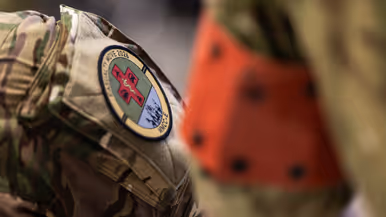 Links: Een militair werkt geconcentreerd achter een bureau met computerschermen. Op de schermen zijn kaarten te zien, terwijl een collega op de achtergrond ook bezig is. Rechts: Een close-up van een militair uniform met een rond embleem ‘Casualty Move 2026’ en een rood kruis. Op de achtergrond is wazig een persoon in oranje zichtbaar.