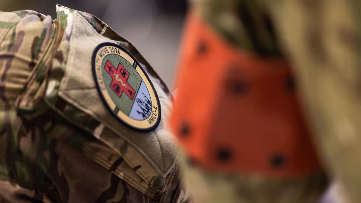 Links: Een militair werkt geconcentreerd achter een bureau met computerschermen. Op de schermen zijn kaarten te zien, terwijl een collega op de achtergrond ook bezig is. Rechts: Een close-up van een militair uniform met een rond embleem ‘Casualty Move 2026’ en een rood kruis. Op de achtergrond is wazig een persoon in oranje zichtbaar.