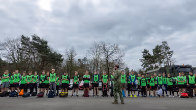 Een groep jongeren in felgroene hesjes met nummers staat in het gelid. Een militair in uniform geeft instructies op een parkeerterrein met bomen en voertuigen.