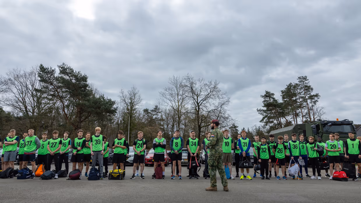 Een groep jongeren in felgroene hesjes met nummers staat in het gelid. Een militair in uniform geeft instructies op een parkeerterrein met bomen en voertuigen.