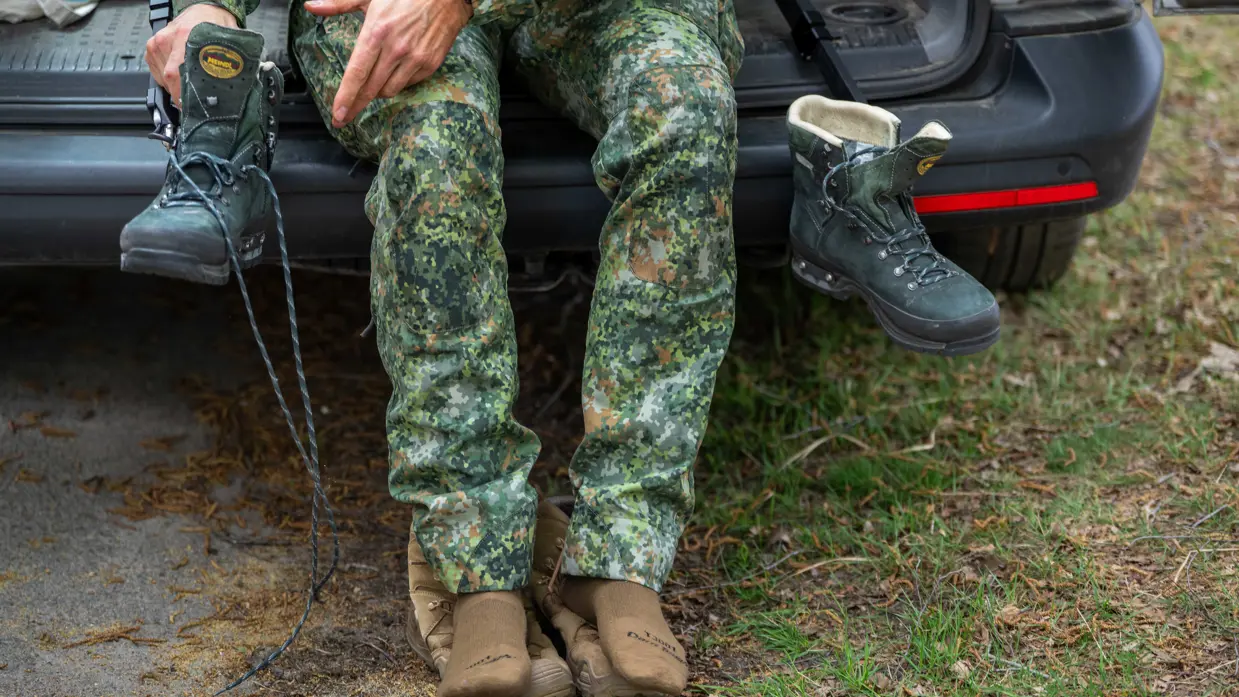 Links: iemand trekt een groene bergschoen aan met een zwarte beugel, buiten op een bosbodem met bladeren.    Midden: Iemand in camouflagebroek zit op een auto met bruine sokken en houdt een groene bergschoen vast. Rechts: Een man in uniform houdt een metertje vast. 
