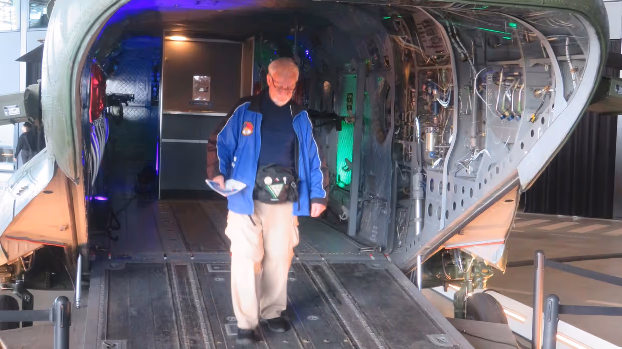 Inkijk in een grote, openstaande militaire helikopter; binnenin zie je stoelen, apparatuur en knipperende groene lichten, terwijl iemand binnen staat.