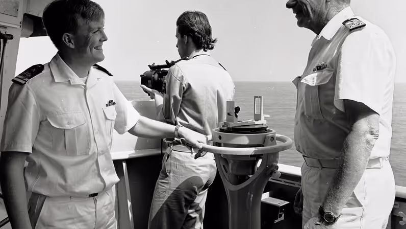 Drie personen in marine-uniform staan op een schip. Twee lachen naar elkaar, één houdt een verrekijker vast. Ze staan bij navigatieapparatuur met zee op de achtergrond.