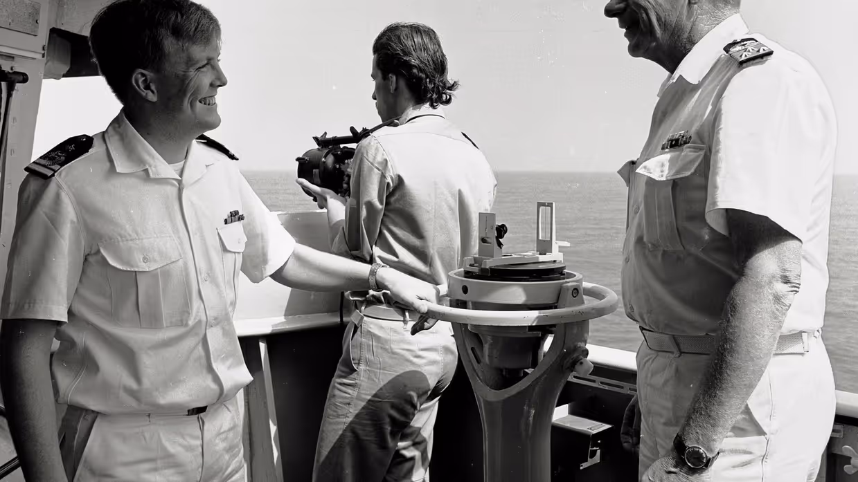 Drie personen in marine-uniform staan op een schip. Twee lachen naar elkaar, één houdt een verrekijker vast. Ze staan bij navigatieapparatuur met zee op de achtergrond.