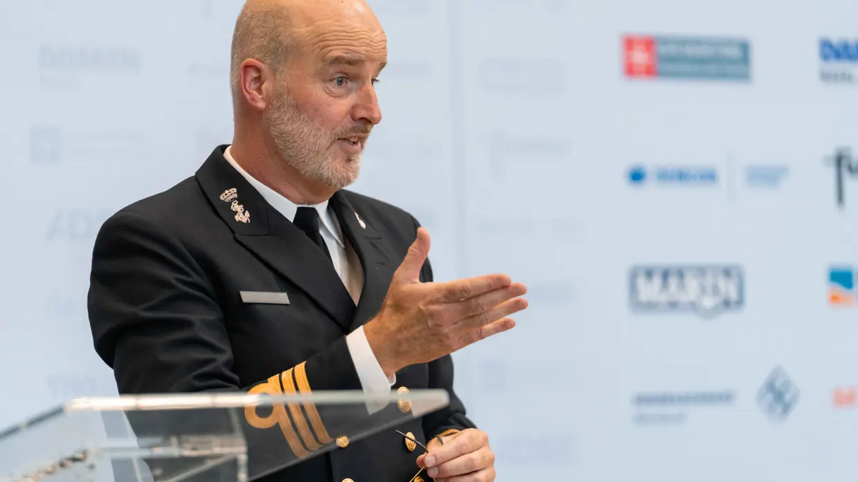 Een kale man in marine-uniform praat en gebruikt daarbij zijn handen.