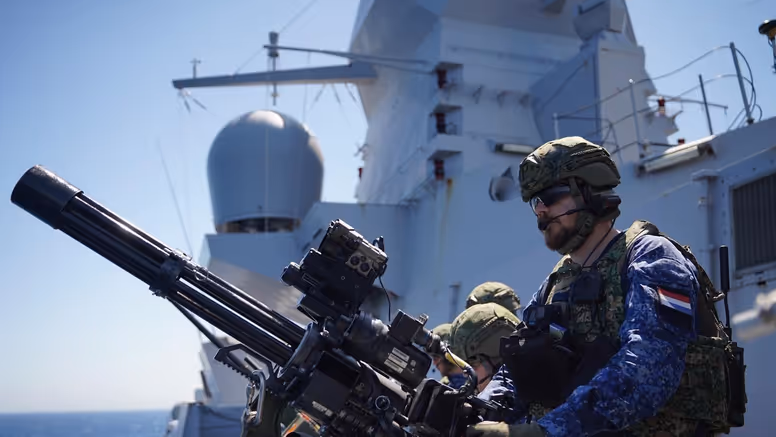 Een bemanningslid met baard staat achter een minigun op een van de dekken van Zr.Ms. De Ruyter