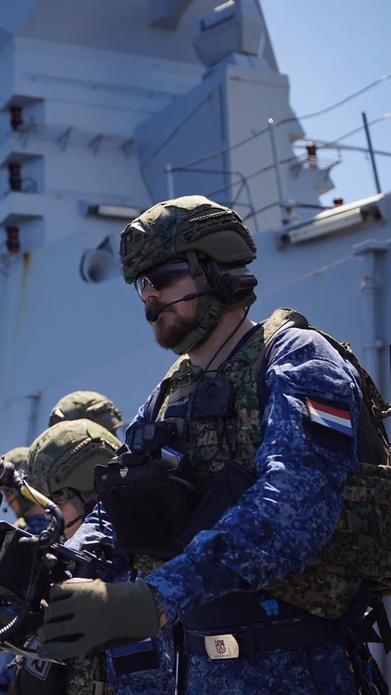 Een bemanningslid met baard staat achter een minigun op een van de dekken van Zr.Ms. De Ruyter