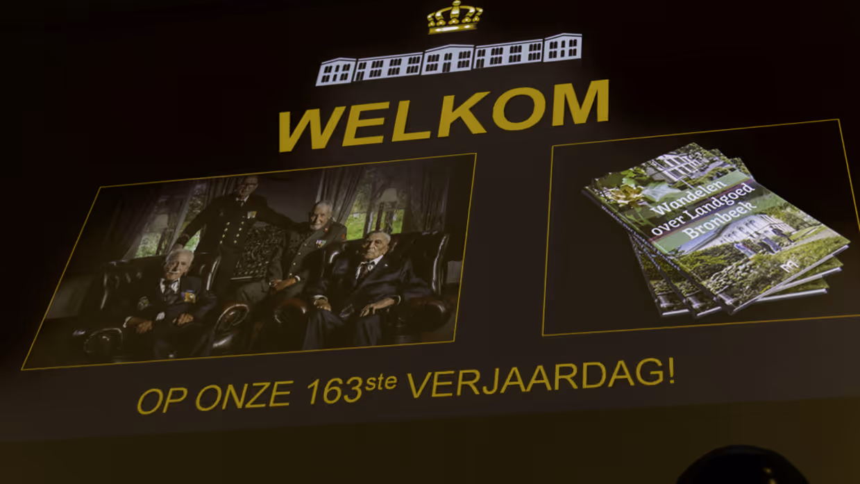 Een groot scherm met daarop een foto van vier  veteranen en een beeld van de wandelgids. Qua tekst staat er: welkom op onze 163e verjaardag. 