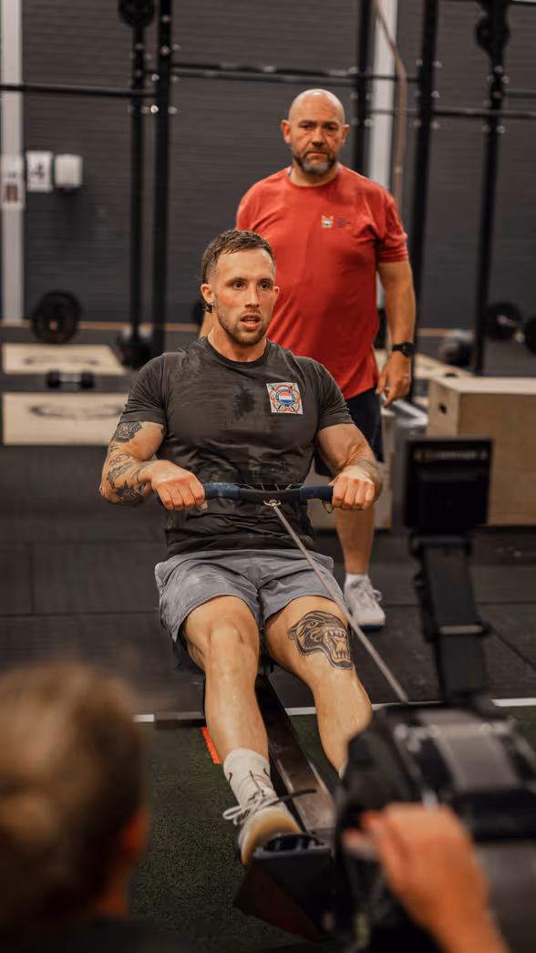 Een man traint op een roeimachine in een sportschool. Hij heeft tatoeages en draagt sportkleding. Een trainer kijkt toe. Apparaat staat centraal.