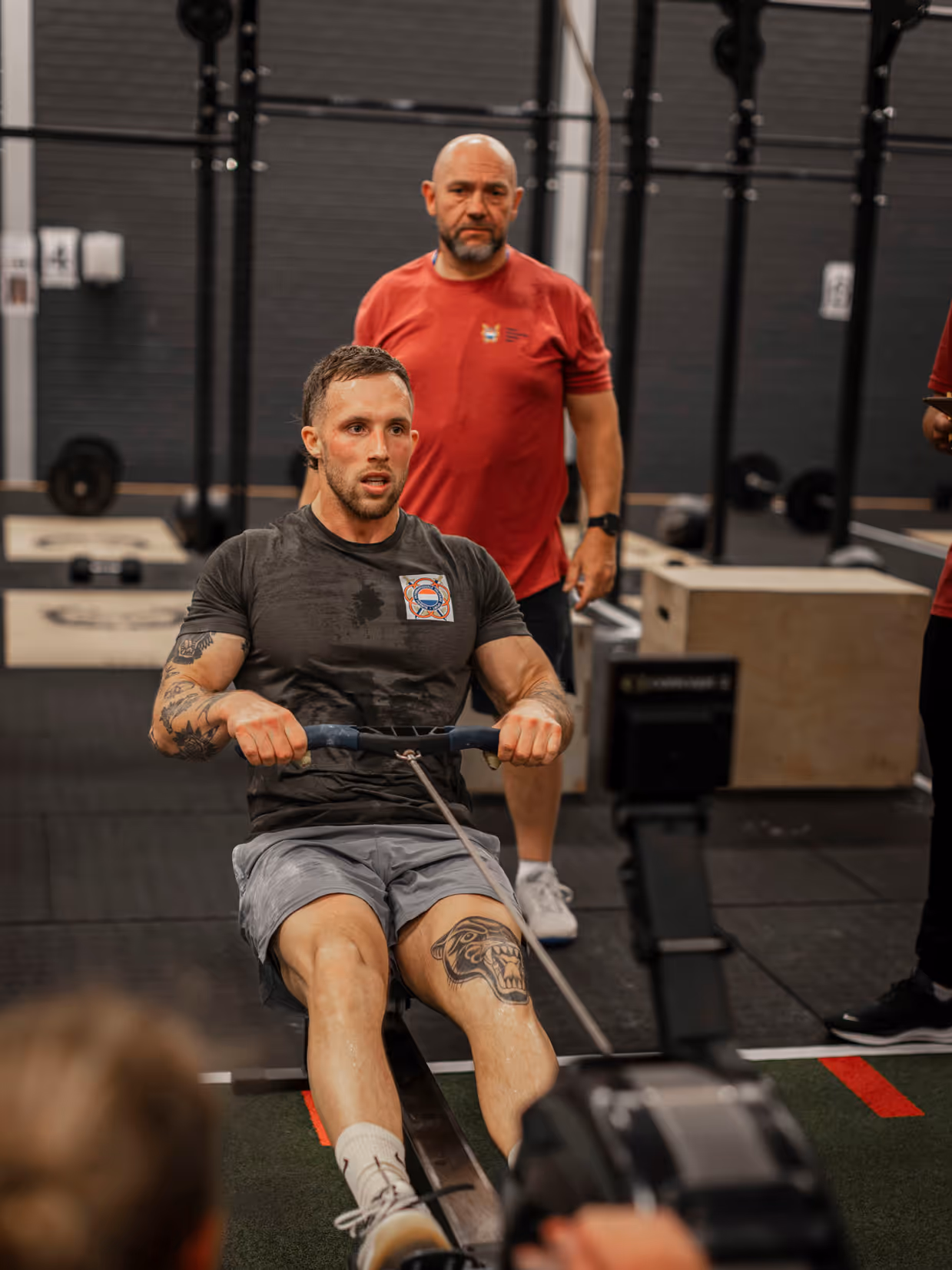 Een man traint op een roeimachine in een sportschool. Hij heeft tatoeages en draagt sportkleding. Een trainer kijkt toe. Apparaat staat centraal.