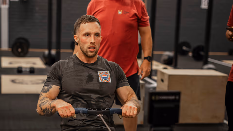 Een man traint op een roeimachine in een sportschool. Hij heeft tatoeages en draagt sportkleding. Een trainer kijkt toe. Apparaat staat centraal.