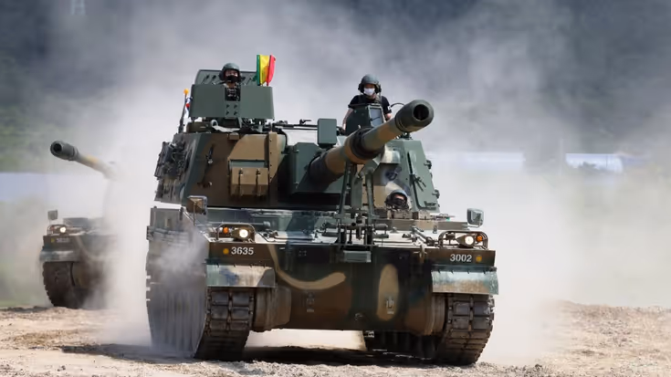 Een zwaar militair voertuig met een kanon rijdt over een stoffig terrein, terwijl een militair boven een luik uitsteekt.