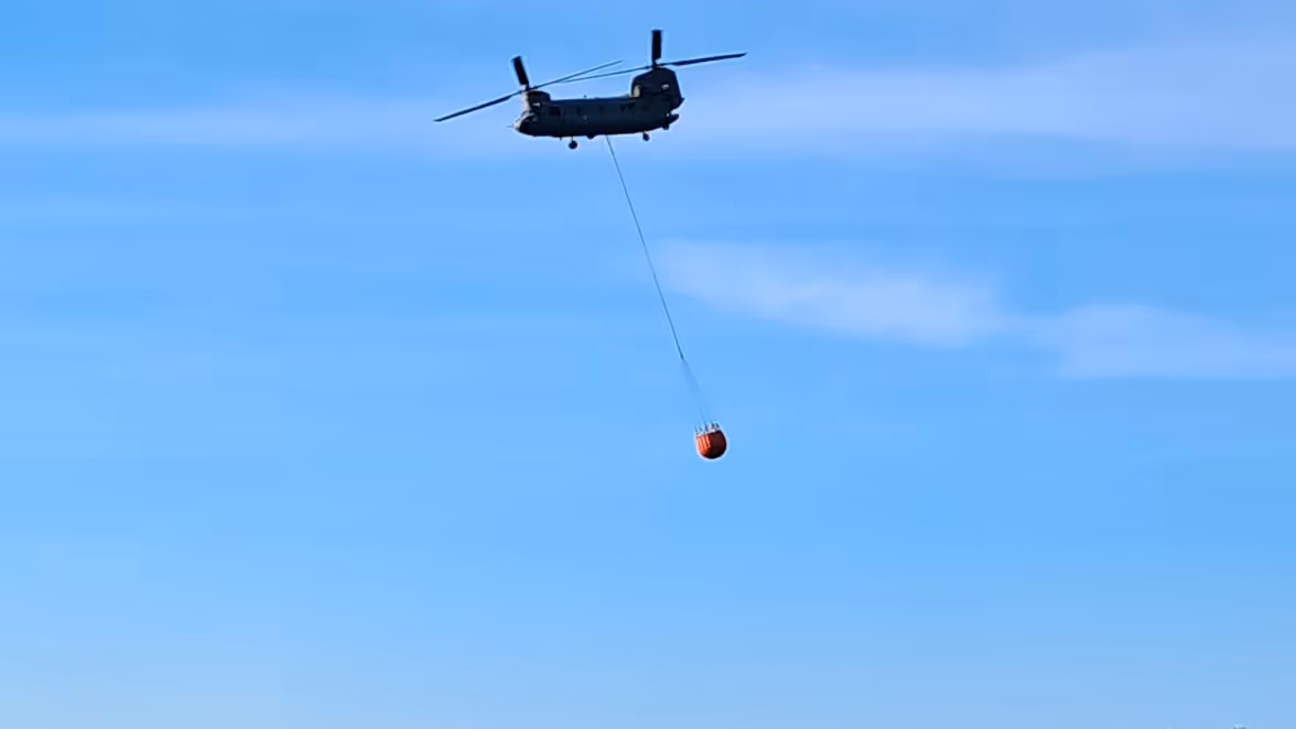 Een grote helikopter vliegt laag boven rustig, blauw water met een lange kabel en een felgekleurde vlag of bal eronder, kustlijn op de achtergrond. 