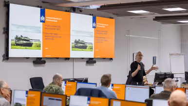 De facilitator van de CDF staat voor de groep en maakt gebruik van zijn handen om de situatie uit te leggen. De aanwezige burgers en militairen luisteren naar hem.