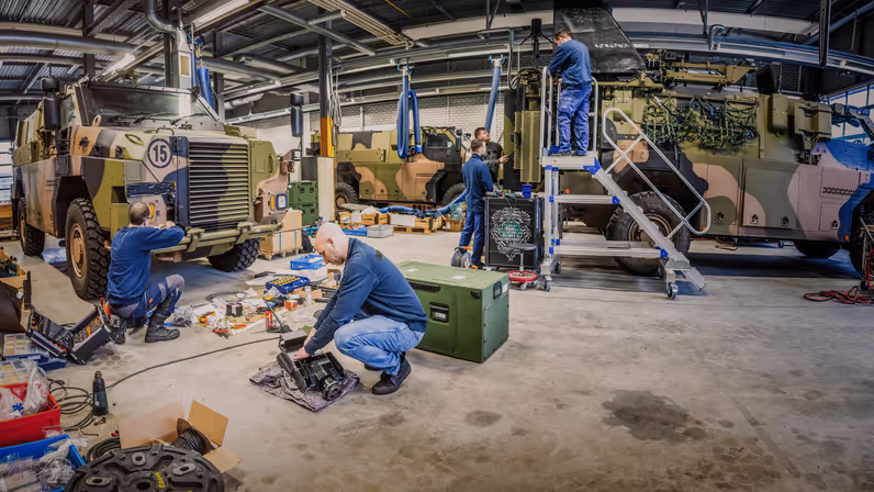 Foto links: een militair boven de Bushmaster, met boven hem de antennes die boven hem uittorenen. Foto rechts: een werkplaats. Hier zijn diverse mensen aan het werk om de Bushmaster om te bouwen voor het bataljon.  