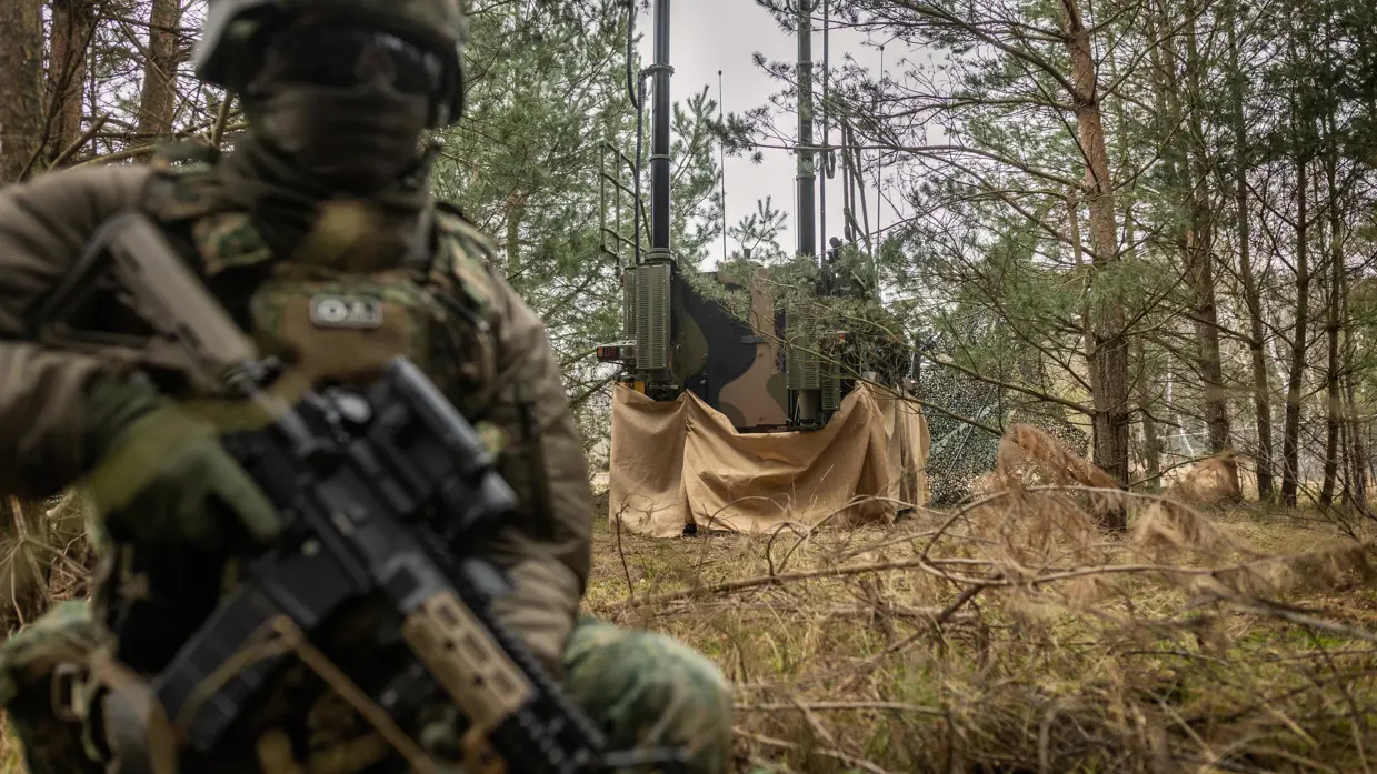 Een bewapende militair zit geknield voor een gecamoufleerd EOV-systeem.