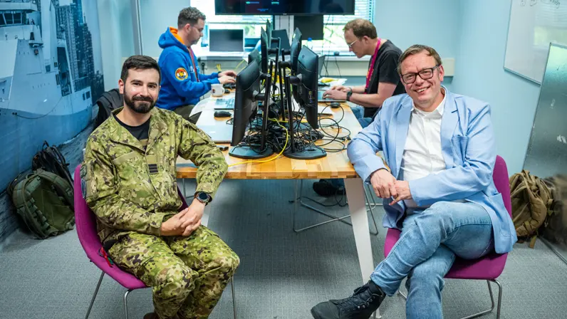 Een burger en een militair poseren op een stoel aan rand van bureau met op achtergrond twee werkende mannen achter een PC.