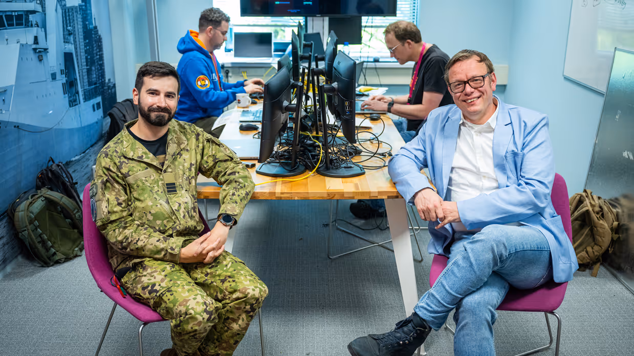 Een burger en een militair poseren op een stoel aan rand van bureau met op achtergrond twee werkende mannen achter een PC.