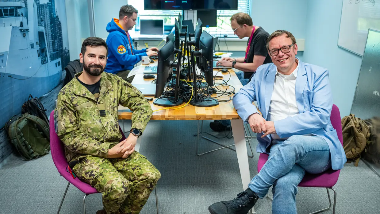 Een burger en een militair poseren op een stoel aan rand van bureau met op achtergrond twee werkende mannen achter een PC.