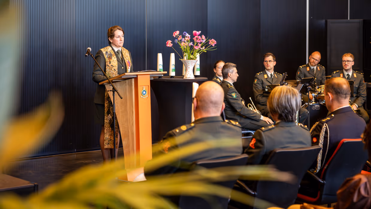 Een militair dominee spreekt de zaal toe. Ze praat door een microfoon. Achter haar zit de militaire muziek. 