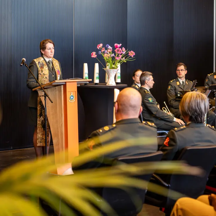 Een militair dominee spreekt de zaal toe. Ze praat door een microfoon. Achter haar zit de militaire muziek. 