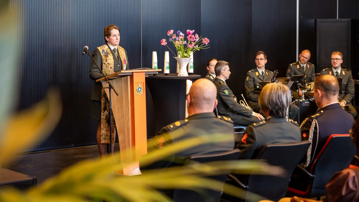Een militair dominee spreekt de zaal toe. Ze praat door een microfoon. Achter haar zit de militaire muziek. 
