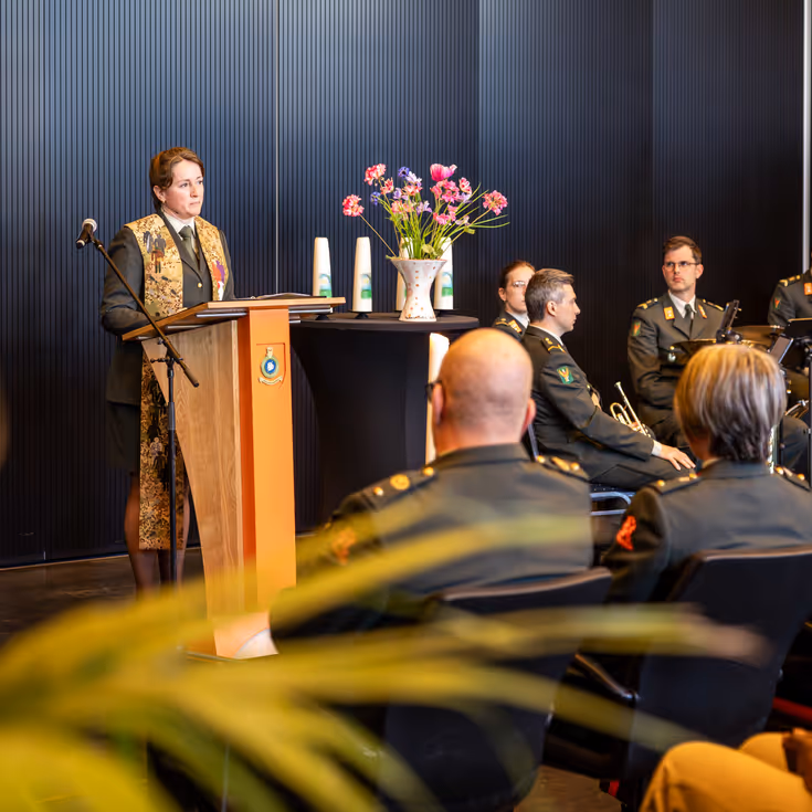 Een militair dominee spreekt de zaal toe. Ze praat door een microfoon. Achter haar zit de militaire muziek. 