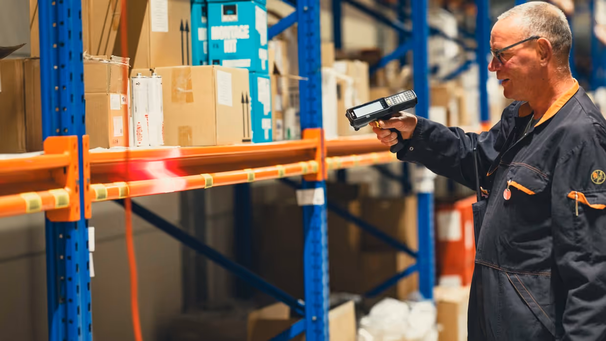  Een man staat voor een magazijnrek. In zijn handen heeft hij een handscanner, waarmee hij een barcode op het rek bliept.