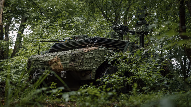 Een militair voertuig vol camouflage houdt zich schuil in een bos. 
