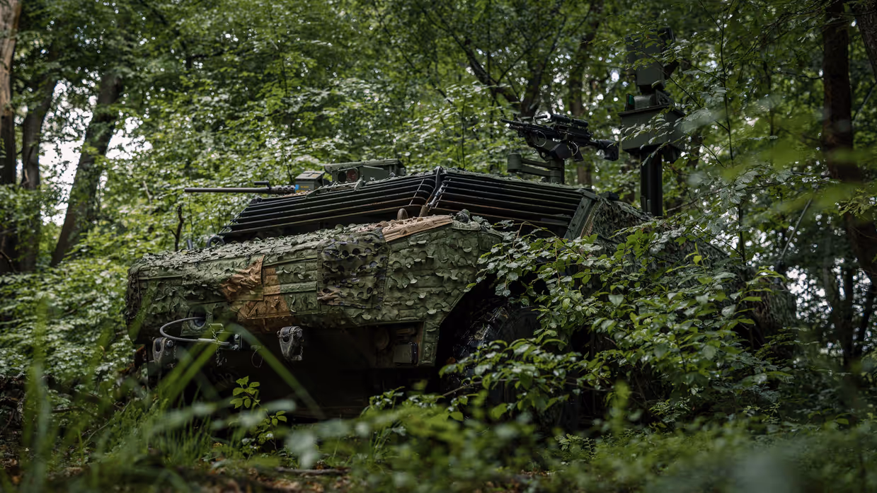 Een militair voertuig vol camouflage houdt zich schuil in een bos. 