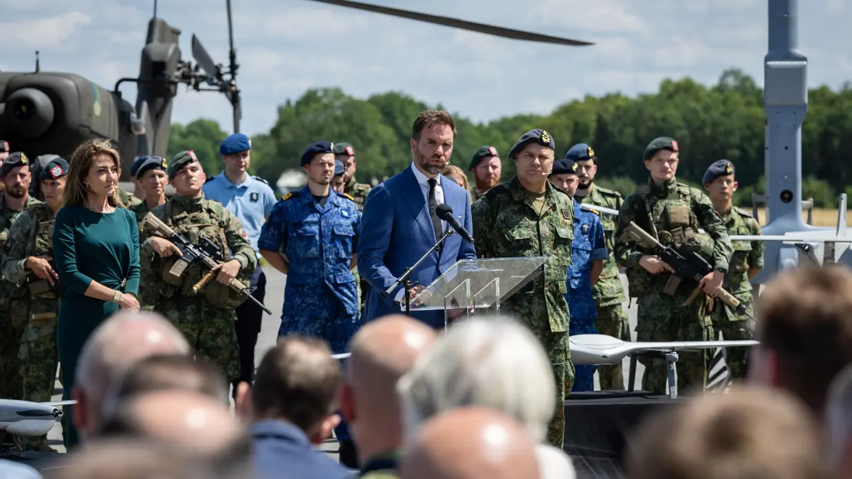 V.l.n.r. de minister, staatssecretaris en Commandant der Strijdkrachten achter drie microfoons. Op de achtergrond staan zowel gewapende als ongewapende militairen opgesteld.