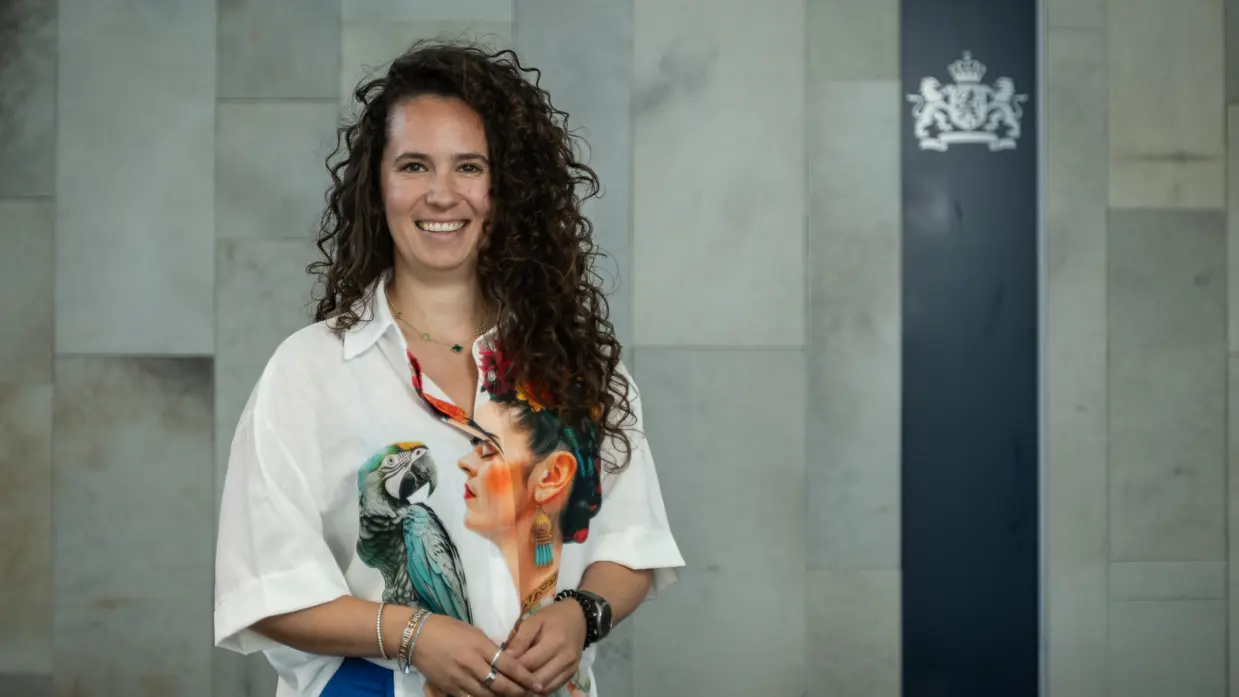 Vrouw met lange, bruine krullen en een wit shirt met afbeeldingen van een papegaai en vrouwenhoofd poseert met haar handen dicht bij elkaar. 