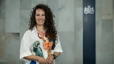 Vrouw met lange, bruine krullen en een wit shirt met afbeeldingen van een papegaai en vrouwenhoofd poseert met haar handen dicht bij elkaar. 