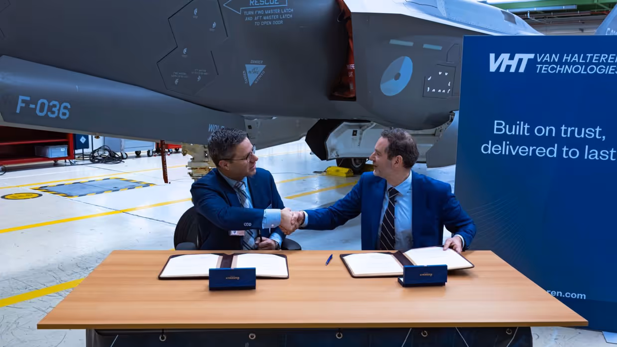 Twee mannen in pak schudden elkaar de hand. Op de tafel waaraan zij zitten liggen contracten. Op de achtergrond staat de replica van de F-35.