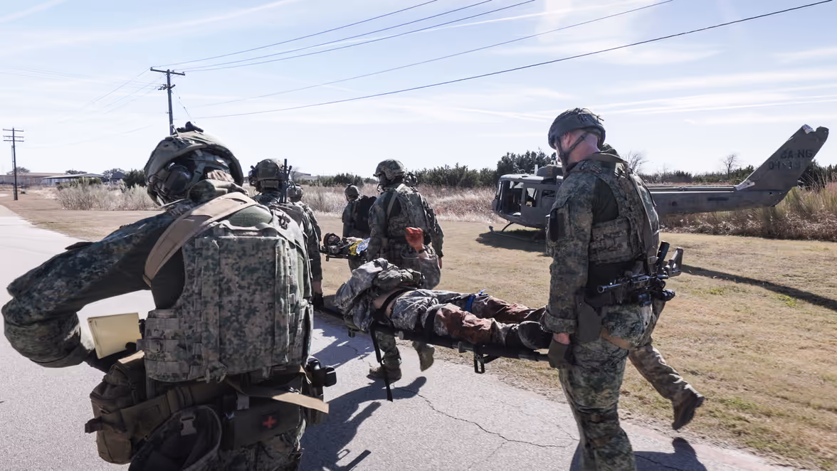  In het veld op een verharde weg dragen militairen een brancard met een gewonde collega.