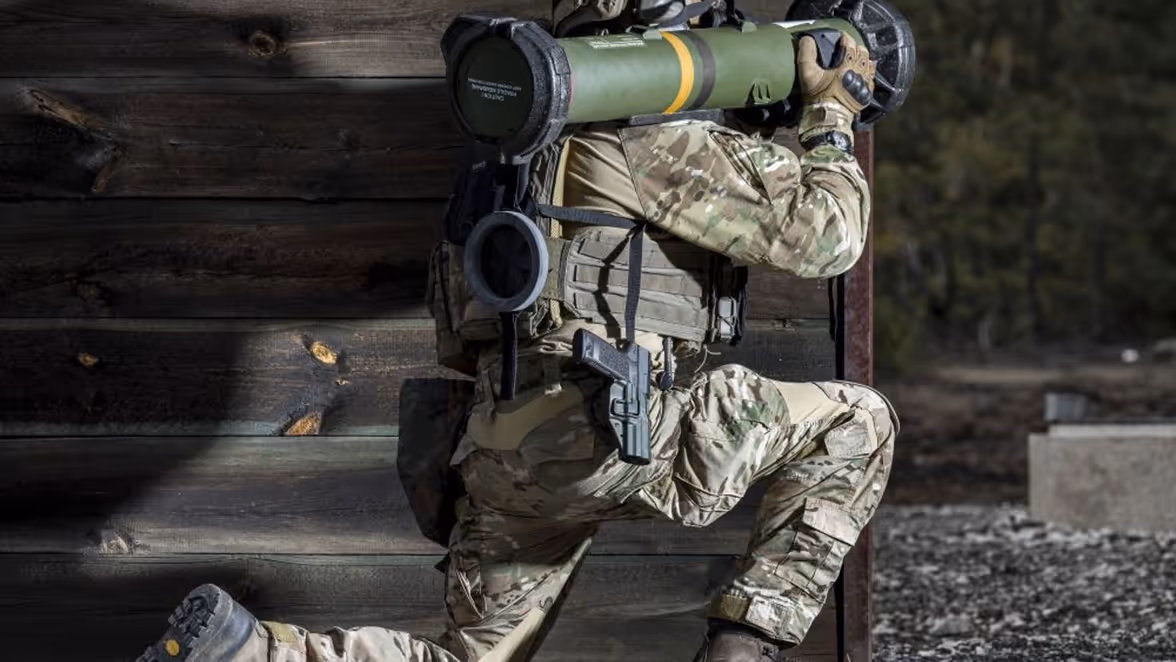 Een militair zit geknield achter een houten muurtje. Op zijn schouder rust het antitankwapen Spike SR.