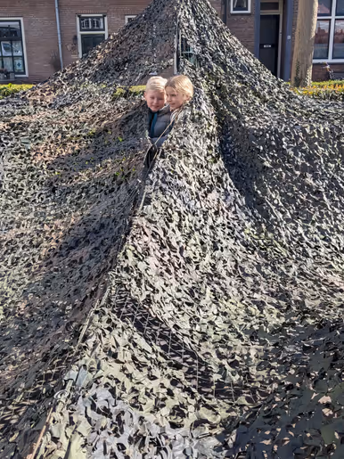Links: Twee glimlachende kinderen kijken onder een grote, groene camouflagenetten-tent vandaan, die buiten voor huizen is opgebouwd en lijkt op een militaire schuilplaats. Rechts: Een glimlachend kind in een militair kogelwerend vest en helm geeft een volwassene in uniform een high-five op een buitenlocatie.