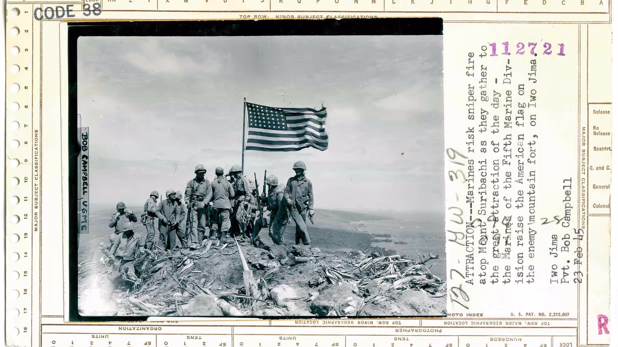 Zes Amerikaanse soldaten hijsen samen de Amerikaanse vlag op een heuveltop tijdens een veldslag. De lucht is bewolkt, de omgeving is rotsachtig en ruig.