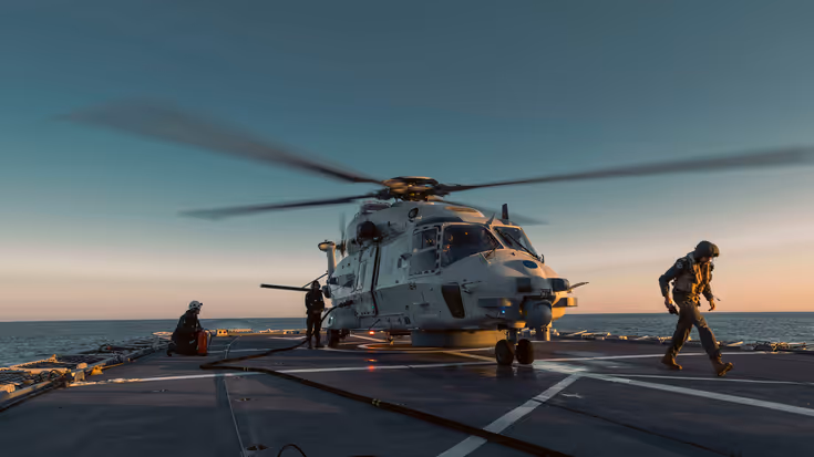 Een grijze militaire helikopter landt op het dek van een schip terwijl twee mannen met helmen op wegrennen.