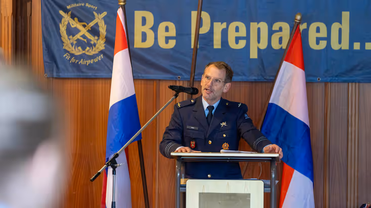 Majoor Twan Relou in uniform achter een spreekgestoelte met een spandoek op de achetrgrond.