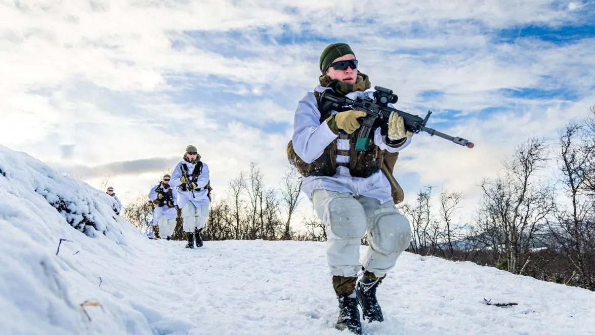 Soldaten in witte wintercamouflage lopen met geweren door een besneeuwd landschap, met een heldere, bewolkte lucht en bomen op de achtergrond.